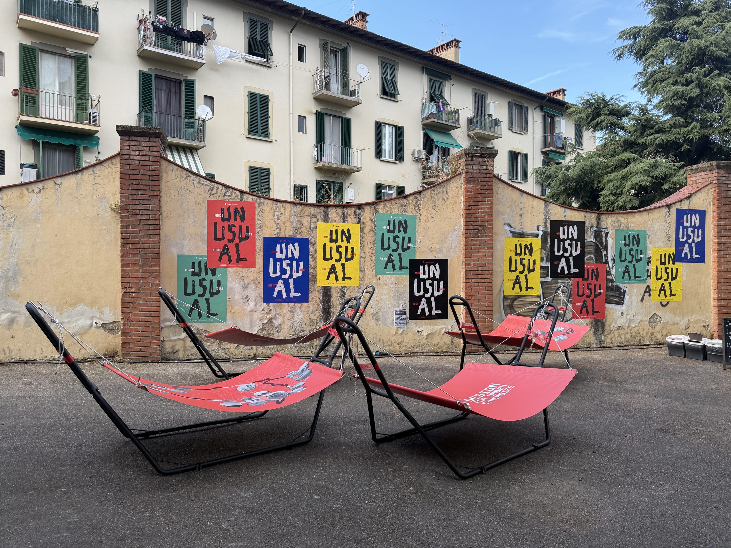 The courtyard at Via Pisana 148: MAP’s red hammocks in front of a yellow wall covered in colourful Unusual posters, with a building with green shutters behind.