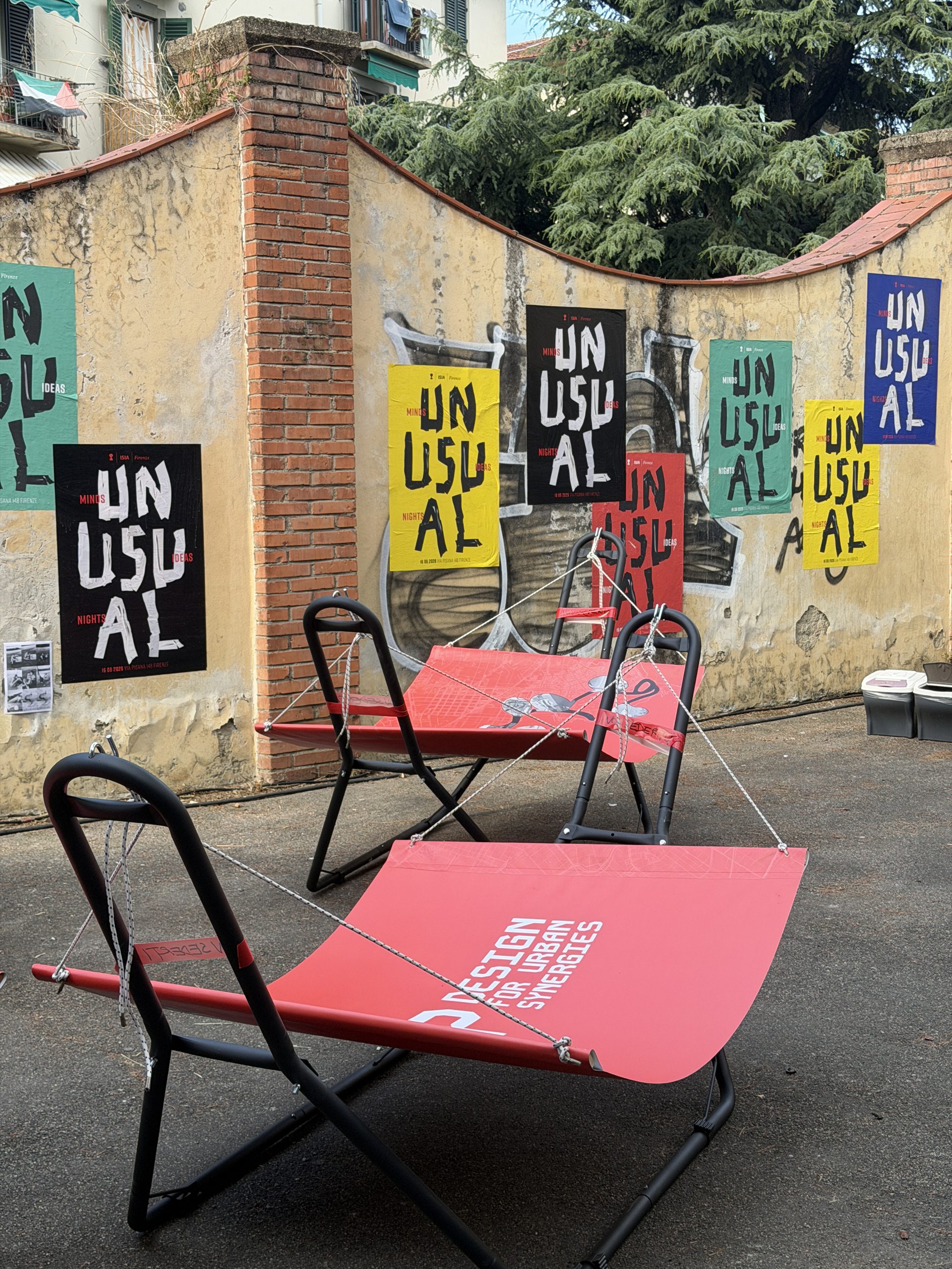 Two red MAP hammocks on black frames; the one in front reads Design for Urban Synergies, with Unusual posters on the wall.