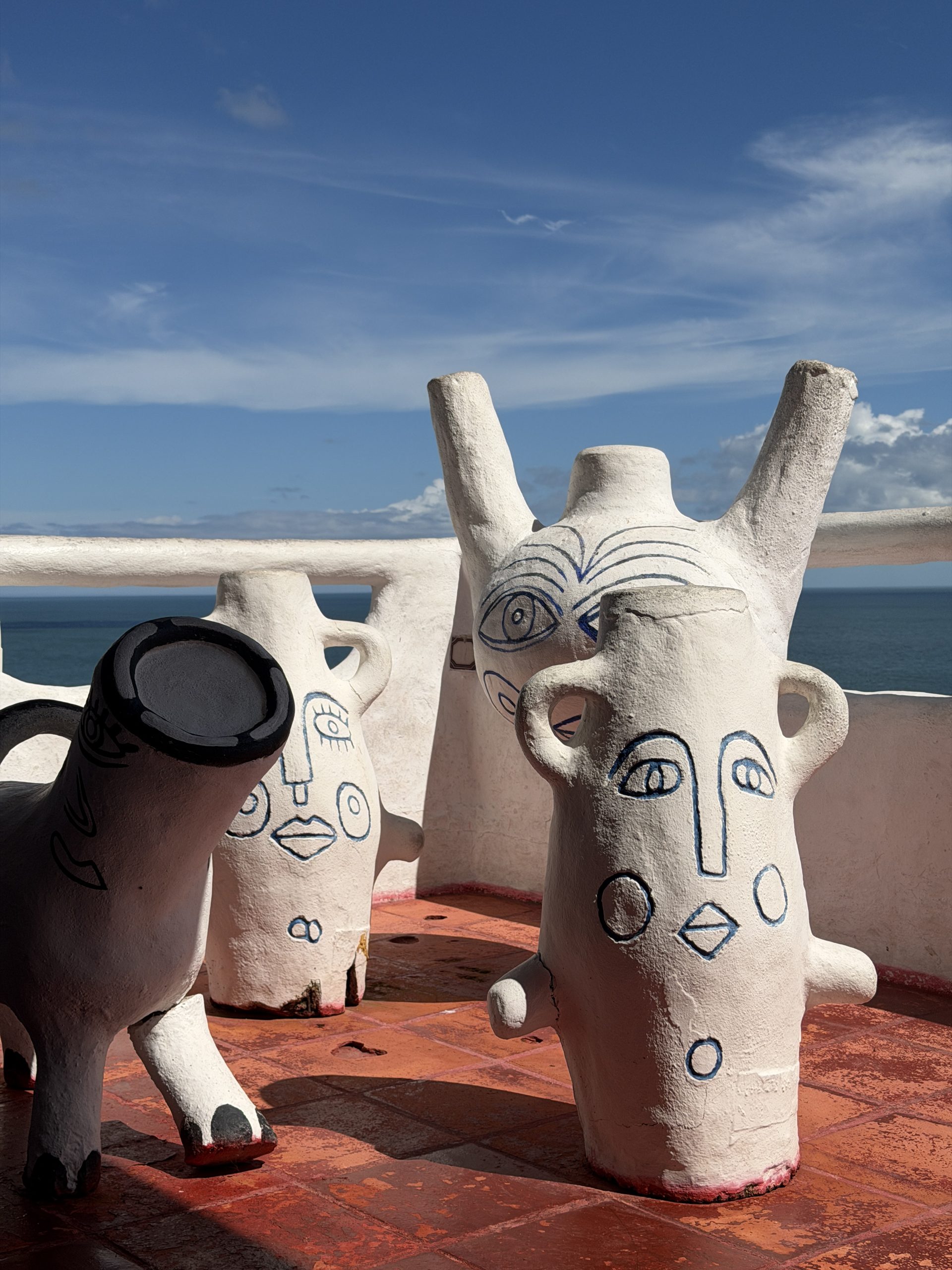 Casapueblo: on a white terrace overlooking the sea, large white ceramic vases with faces drawn in blue and a black and white animal, under a blue sky.