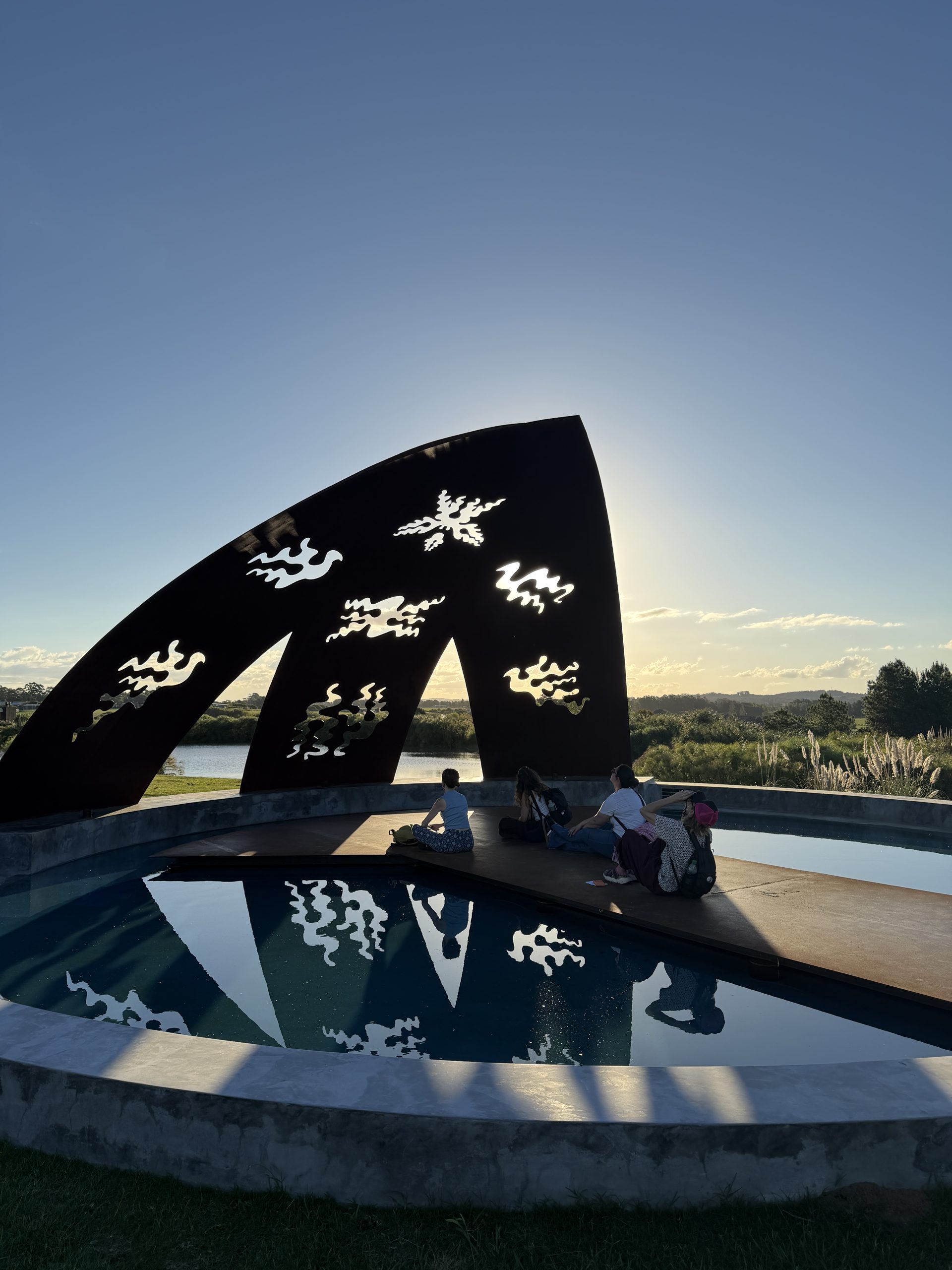 At MACA, towards sunset: a large dark sculpture cut with flame-like shapes above a pool that reflects it; a few people sitting on the wooden platform in the middle.