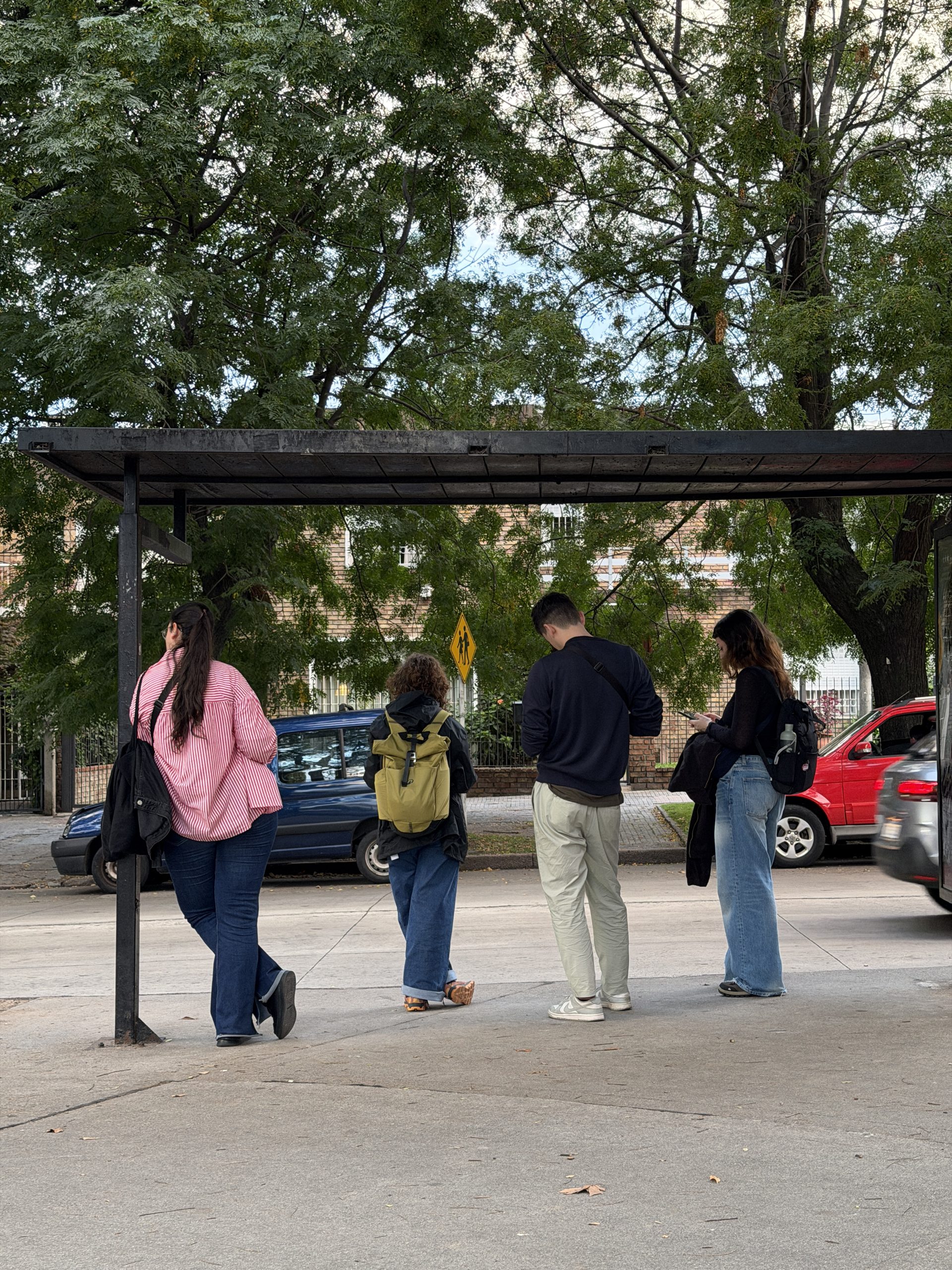 Four of us seen from behind under a bus shelter on a tree-lined street in Montevideo: backpacks, jeans, someone checking their phone.