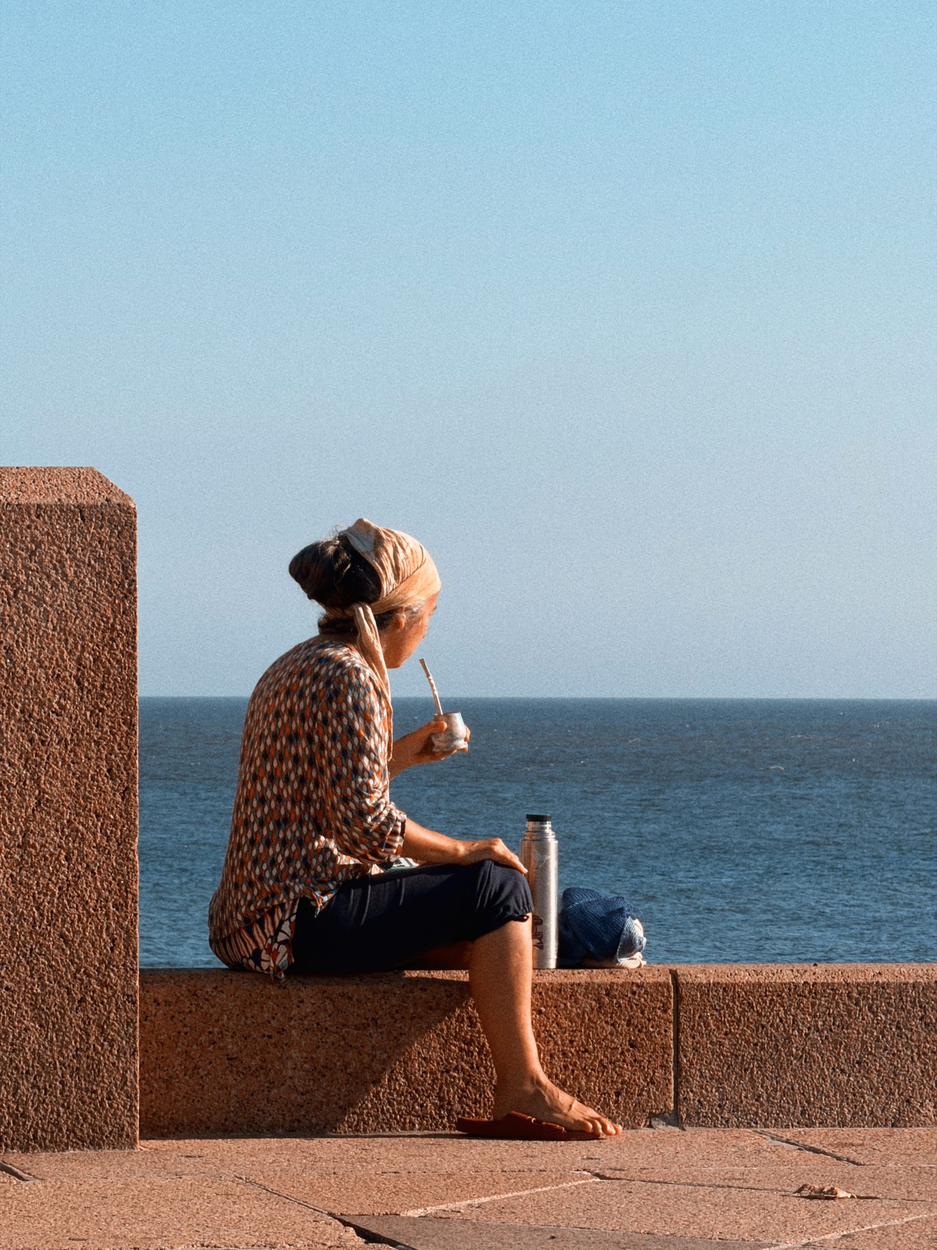 A woman in a headscarf sits on the red granite wall of the rambla drinking mate, a thermos beside her, the Río de la Plata in front.